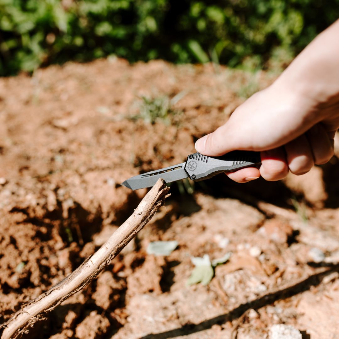 A close-up of a hand holding an otf knife with a silver blade and black handle, positioned over a dry twig in an outdoor dirt setting.