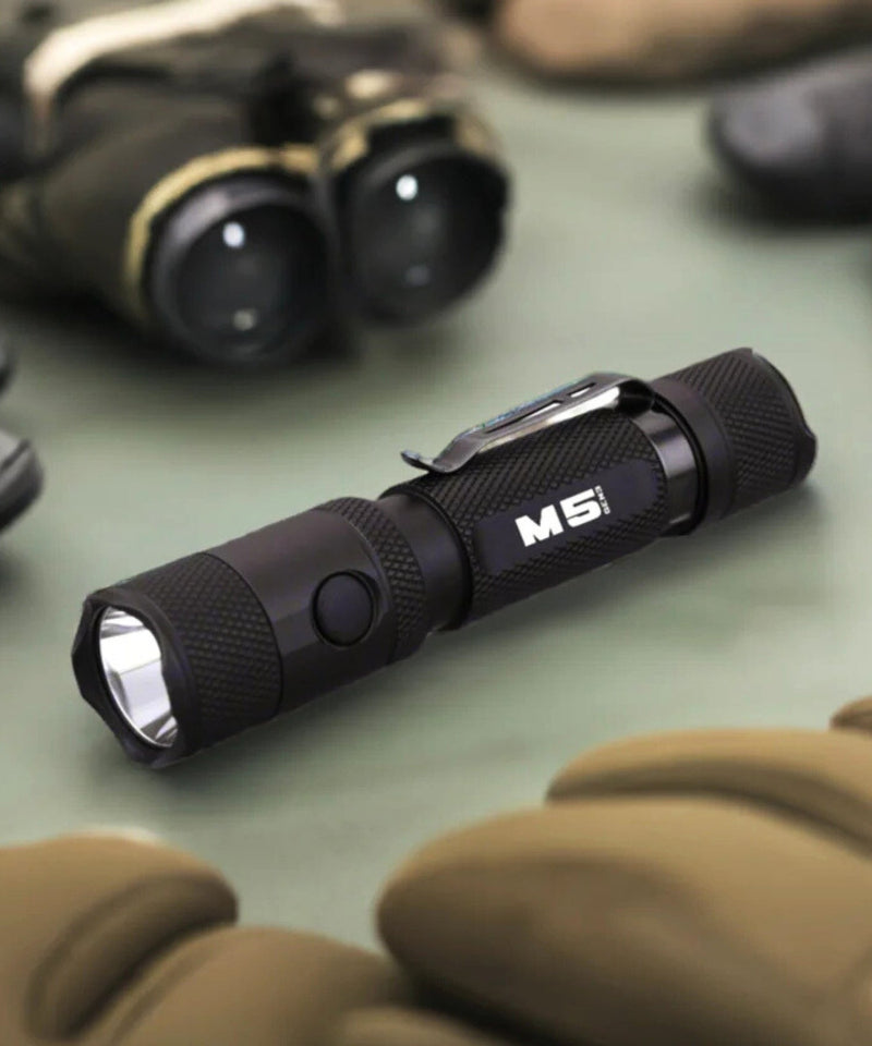Black flashlight with 'M5' branding on a green surface with binoculars and gloves in the background.